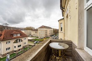 A balcony with a table and chairs, overlooking a street with buildings and parked cars.