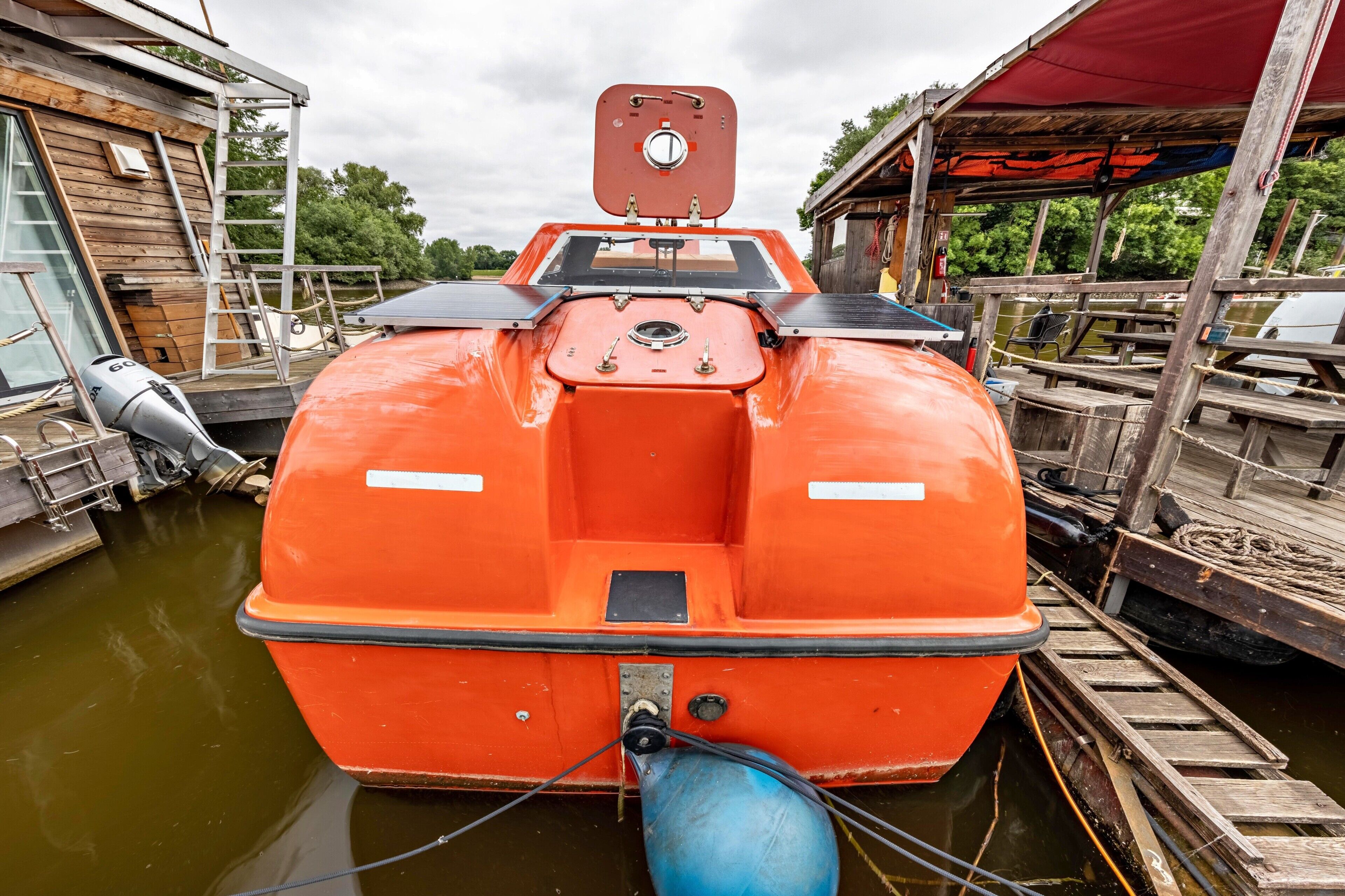 A stationary orange boat docked at a pier with a ladder, a winch, and a covered area with a red canopy.