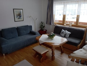Un salon avec un canapé bleu, une table en bois recouverte d’une nappe blanche, un banc et une fenêtre avec des rideaux.