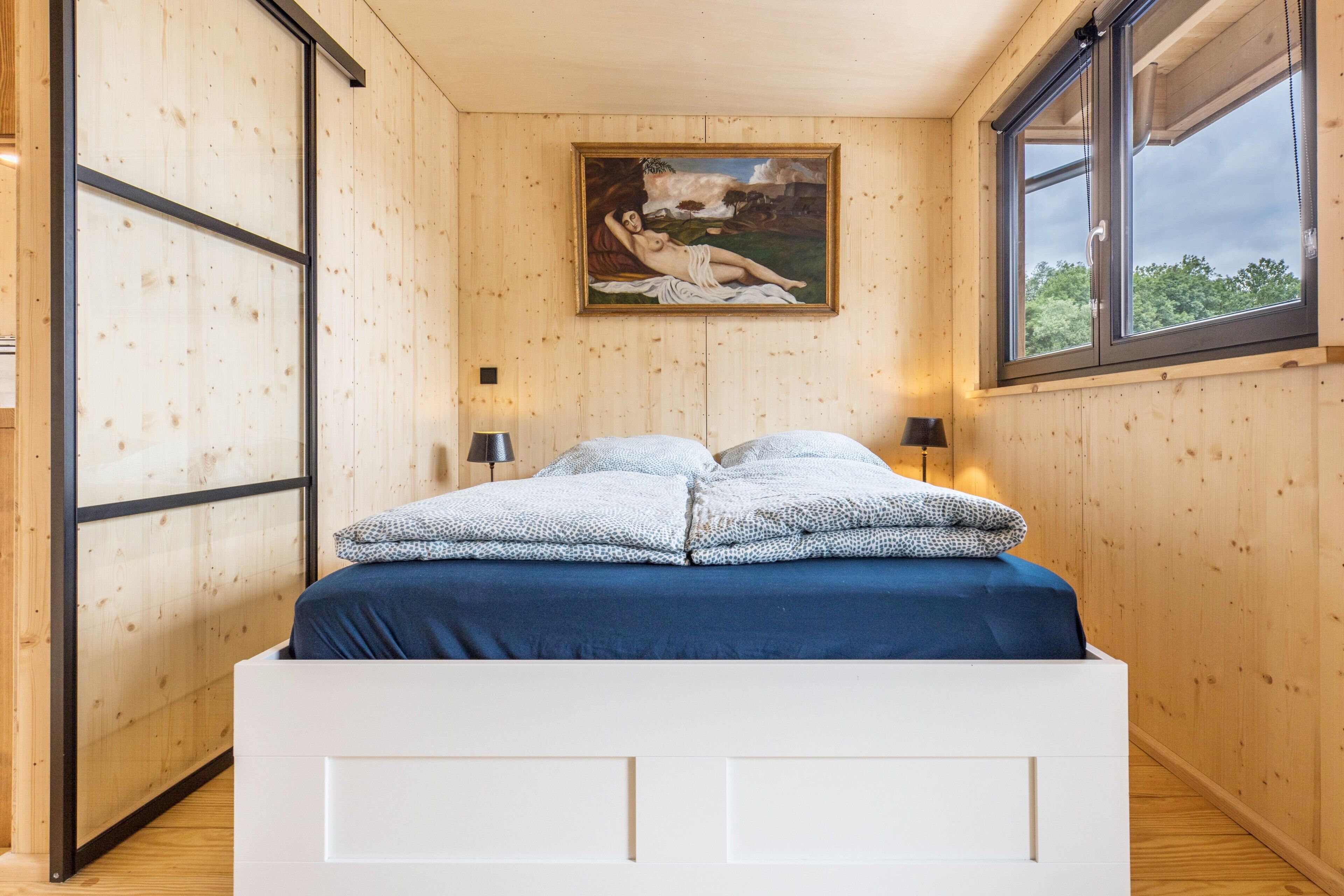 Ein Zimmer mit Holzwänden, einem Bett, einem Gemälde und einem Fenster.