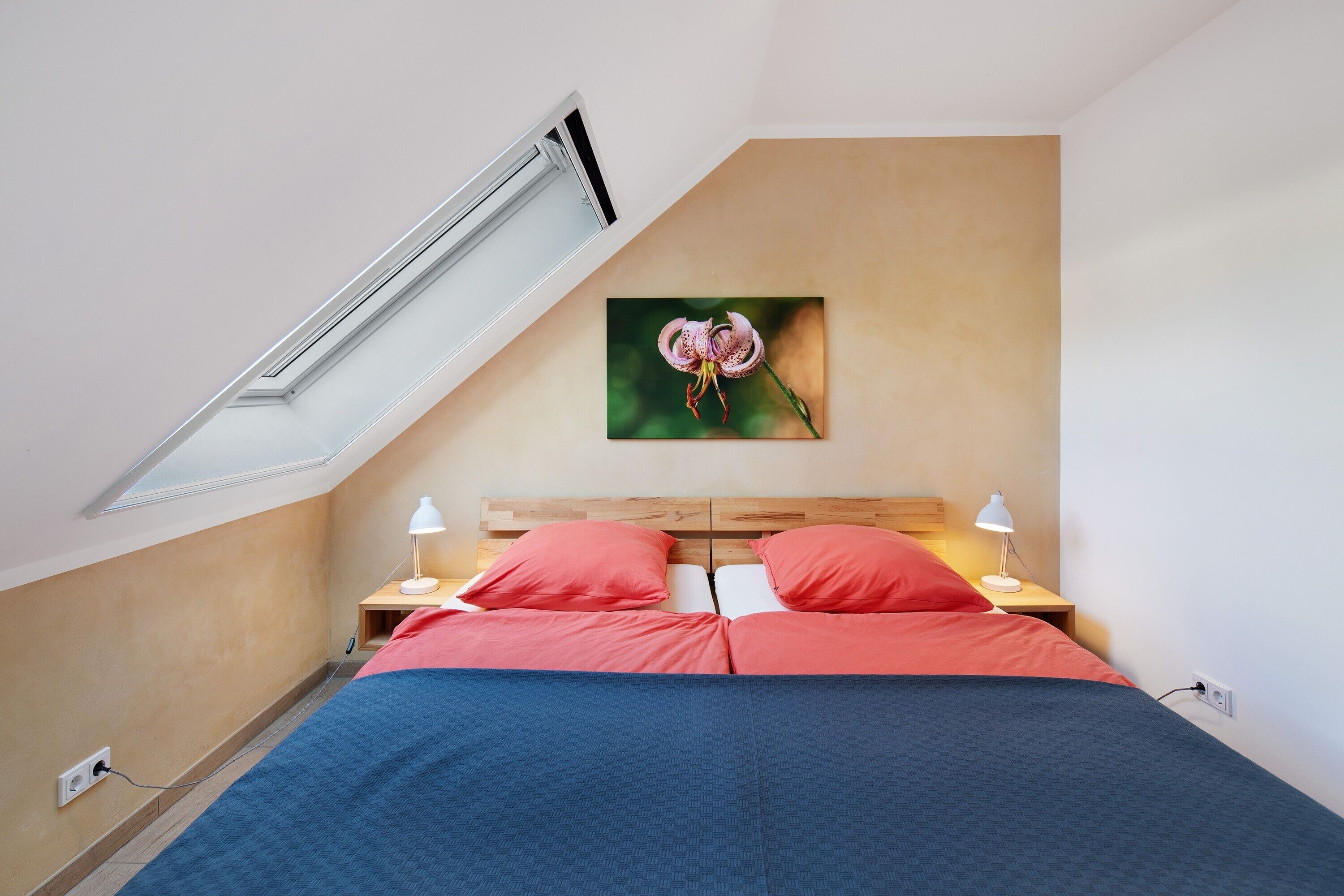 Une chambre avec un plafond en pente, un lit avec une literie rouge, une tête de lit en bois, deux lampes de chevet et un tableau représentant une fleur accroché au mur.