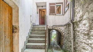 Una estrecha escalera de piedra que lleva a una puerta de madera, con una pared de piedra rústica a un lado y una ventana con persianas al otro.