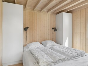 Un dormitorio con paredes de madera, una cama con ropa blanca, dos lámparas fijadas a la pared y un armario blanco.
