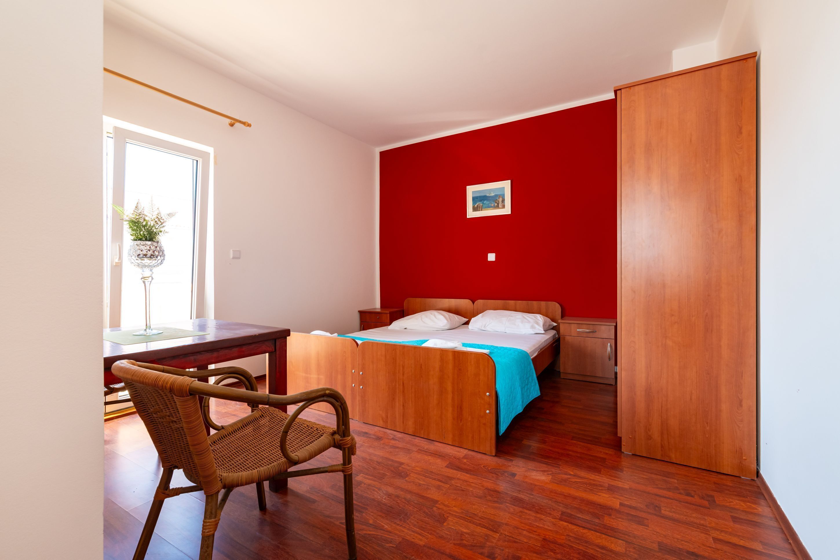 Een slaapkamer met een bed, een houten garderobe, een bureau met stoel, en een raam met een plant.