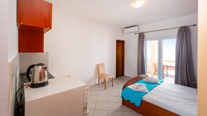A compact room with a kitchenette, a bed with a blue blanket, a chair, and a view of the outdoors through a door.