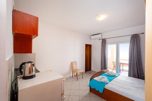Ein kleines Zimmer mit einer KĂŒchenzeile, einem Bett mit blauer Decke, einem Stuhl und Blick ins Freie durch eine TĂŒr.