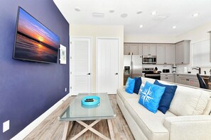 A modern living room with a glass coffee table, a sofa with blue cushions, a flat-screen TV on the wall, and a kitchen area with stainless steel appliances.