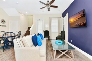 A living room with a sofa, chairs, a glass coffee table, a ceiling fan, and a painting on the wall.