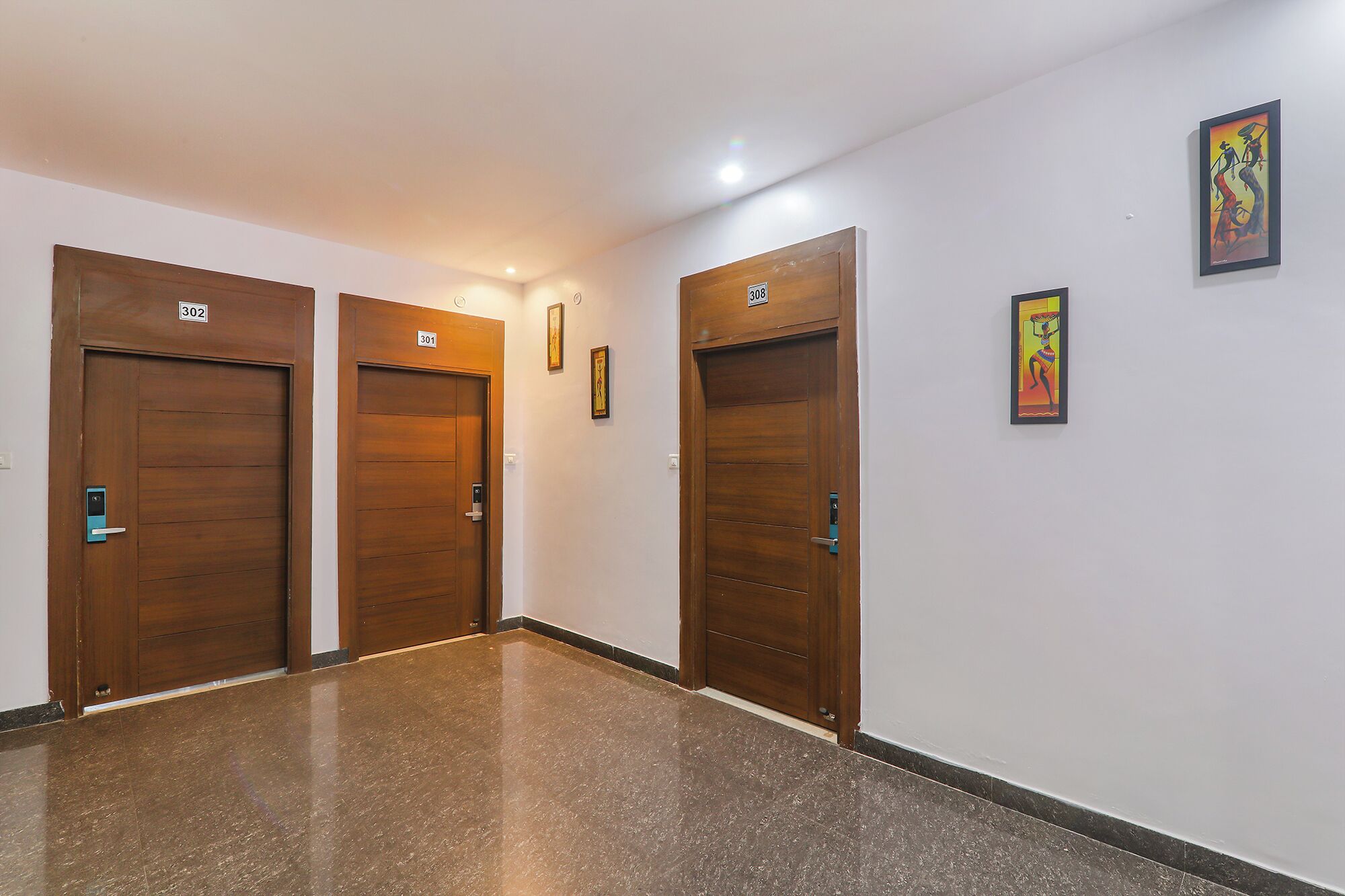 Un couloir avec trois portes en bois, chacune portant un numéro. Le sol est recouvert de marbre poli, et deux œuvres d’art encadrées ornent les murs.