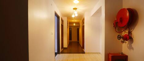 A hallway with a wooden floor, a red fire extinguisher mounted on the wall, and a light fixture hanging from the ceiling.