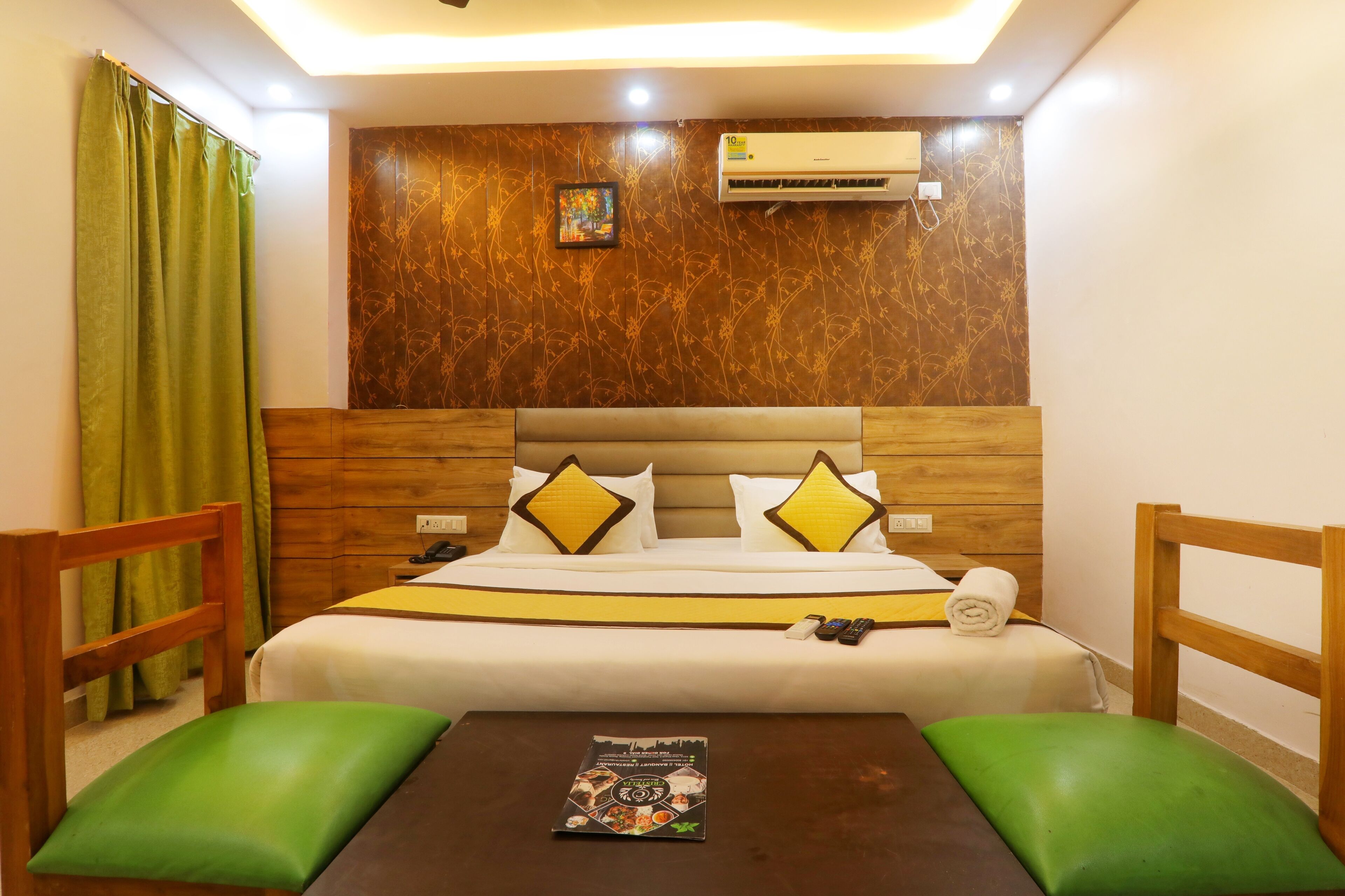 A hotel room with a wooden headboard, a bed with white and yellow bedding, a green chair, a small table with a book, and an air conditioning unit on the wall.