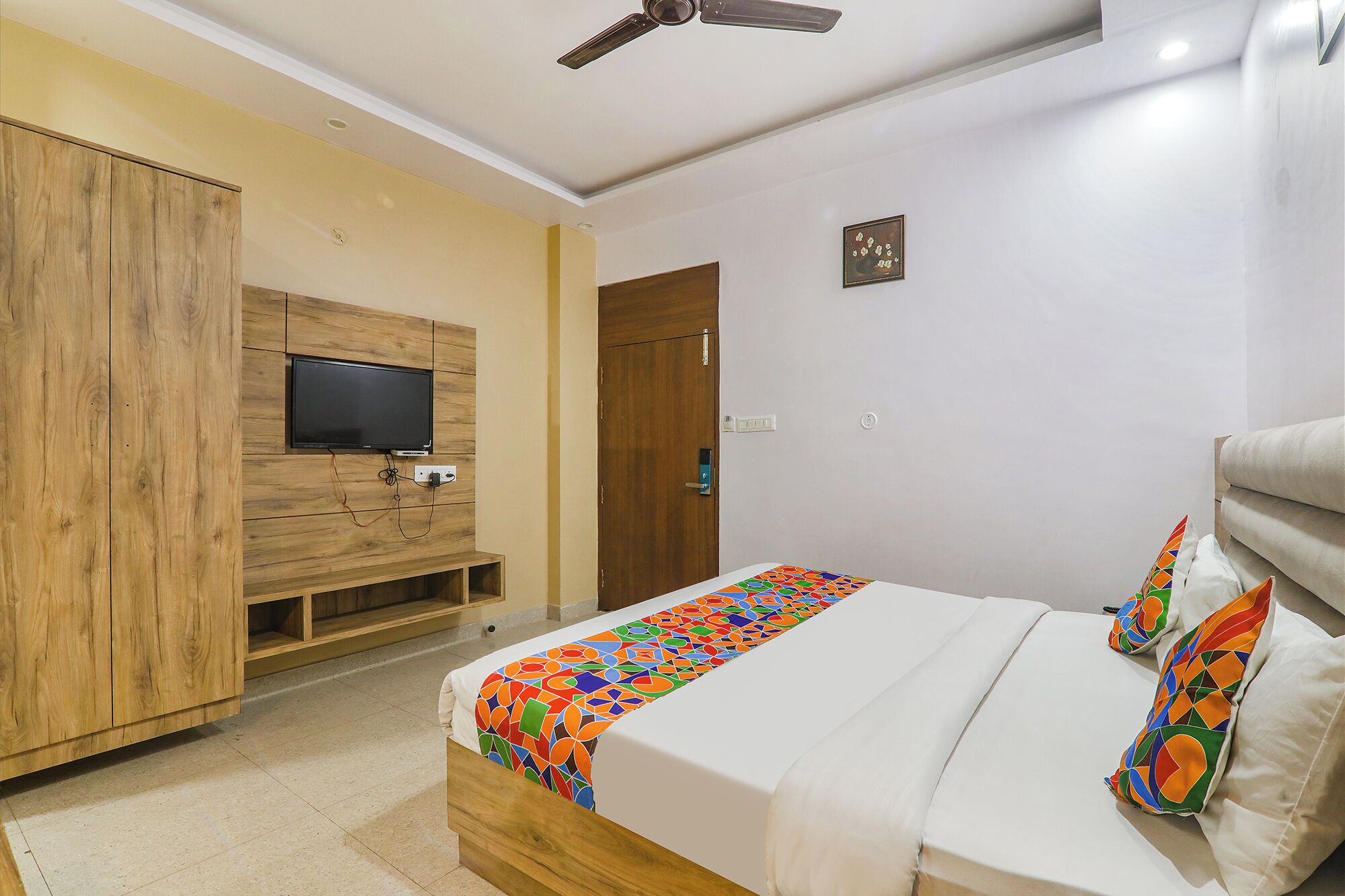 A bedroom with a bed, a TV mounted on a wooden panel, a door, and a ceiling fan.