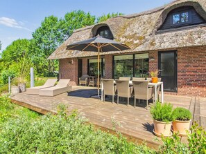 Een huis met rieten dak, een houten terras, buitenmeubels en potplanten.