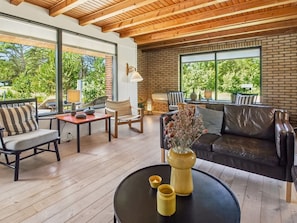 A modern living room with a wooden ceiling, large windows, a sofa, chairs, a table, and a vase with flowers.