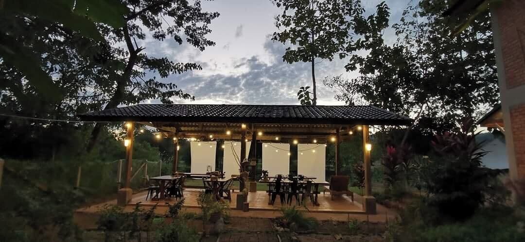 A covered outdoor seating area with tables and chairs, surrounded by trees and a brick wall.