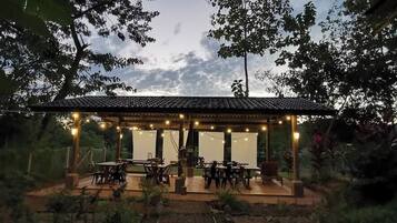 A covered outdoor seating area with tables and chairs, surrounded by trees and a brick wall.