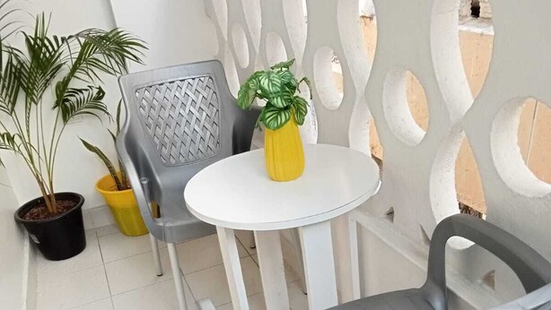 A balcony with two grey plastic chairs, a small white table, and a yellow vase with a plant. There are potted plants on the floor and a white decorative fence with cut-out shapes. - Lux Suites Vee Studio Apartments Bamburi (Mombasa)