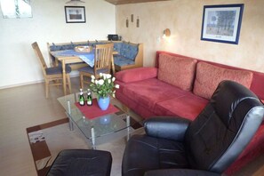 A living room with a glass coffee table, a red sofa, a black leather armchair, and a dining area with a blue tablecloth and wooden chairs.