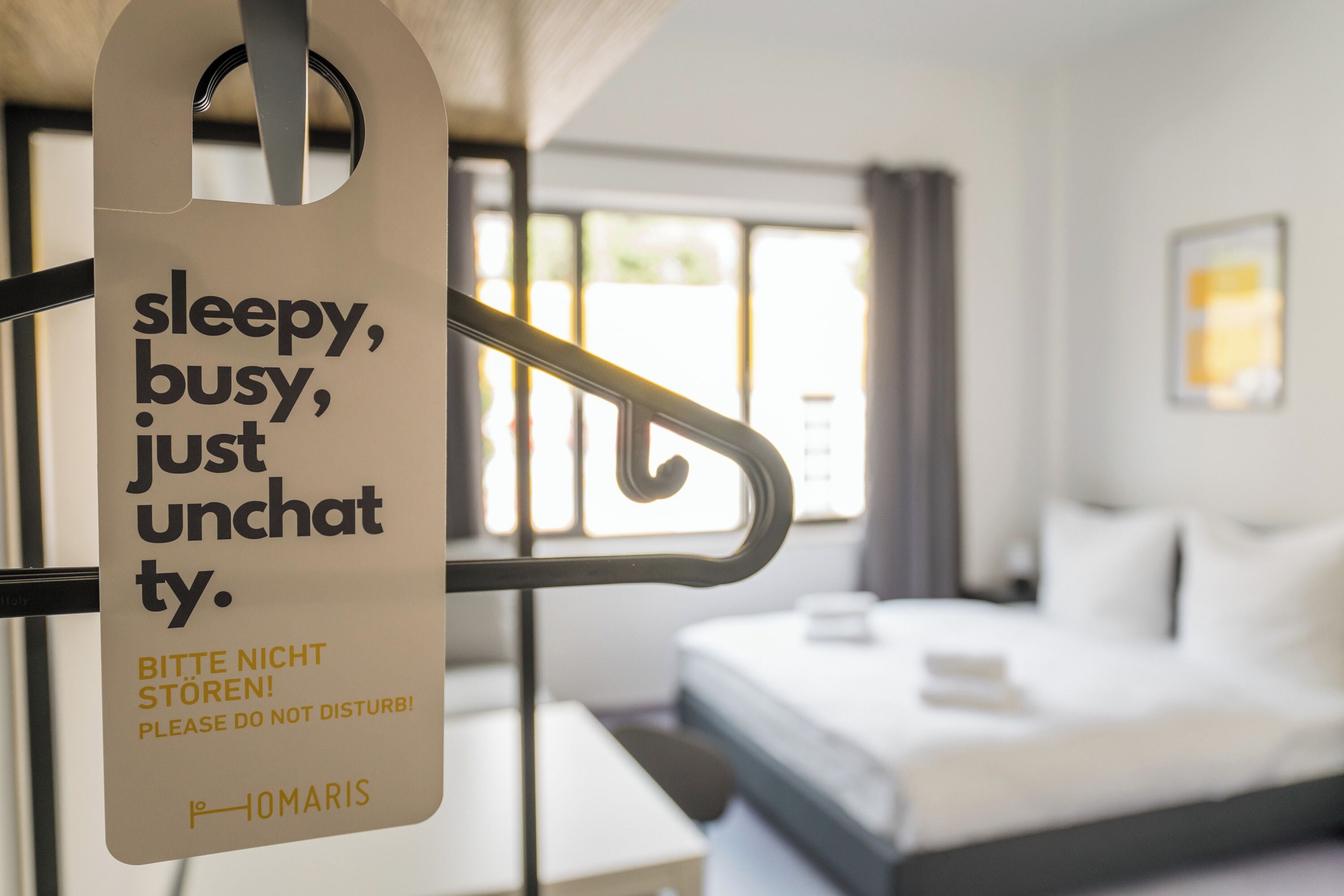 Une chambre d’hôtel avec un lit, un bureau et une fenêtre. Une pancarte sur la porte indique : « Ne pas déranger, je suis occupé(e), je ne suis pas d’humeur à discuter. »