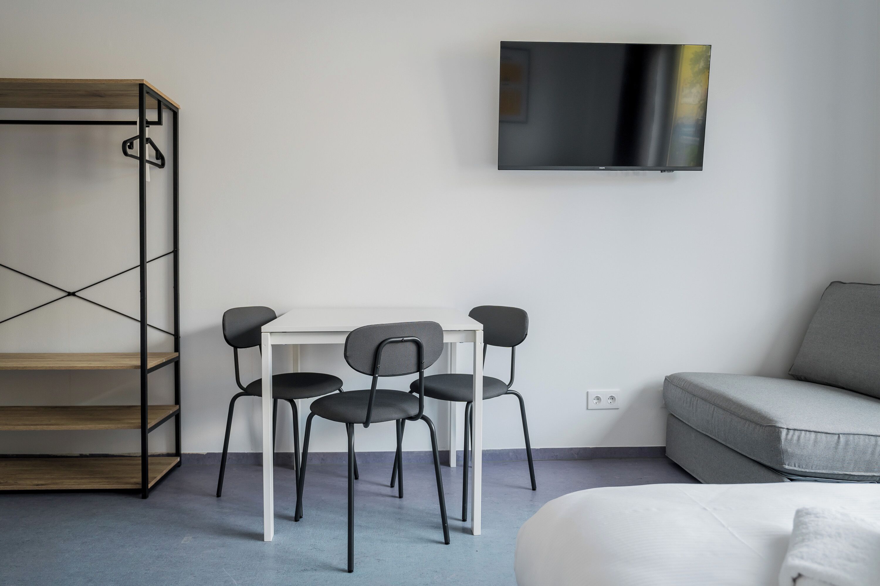 A modern room with a white sofa, a TV mounted on the wall, a desk with two chairs, and a metal shelving unit.