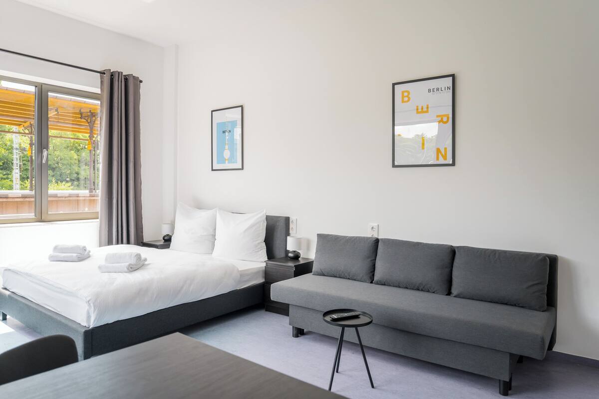 A modern hotel room with a bed, a grey sofa, a small table, and two framed pictures on the wall.