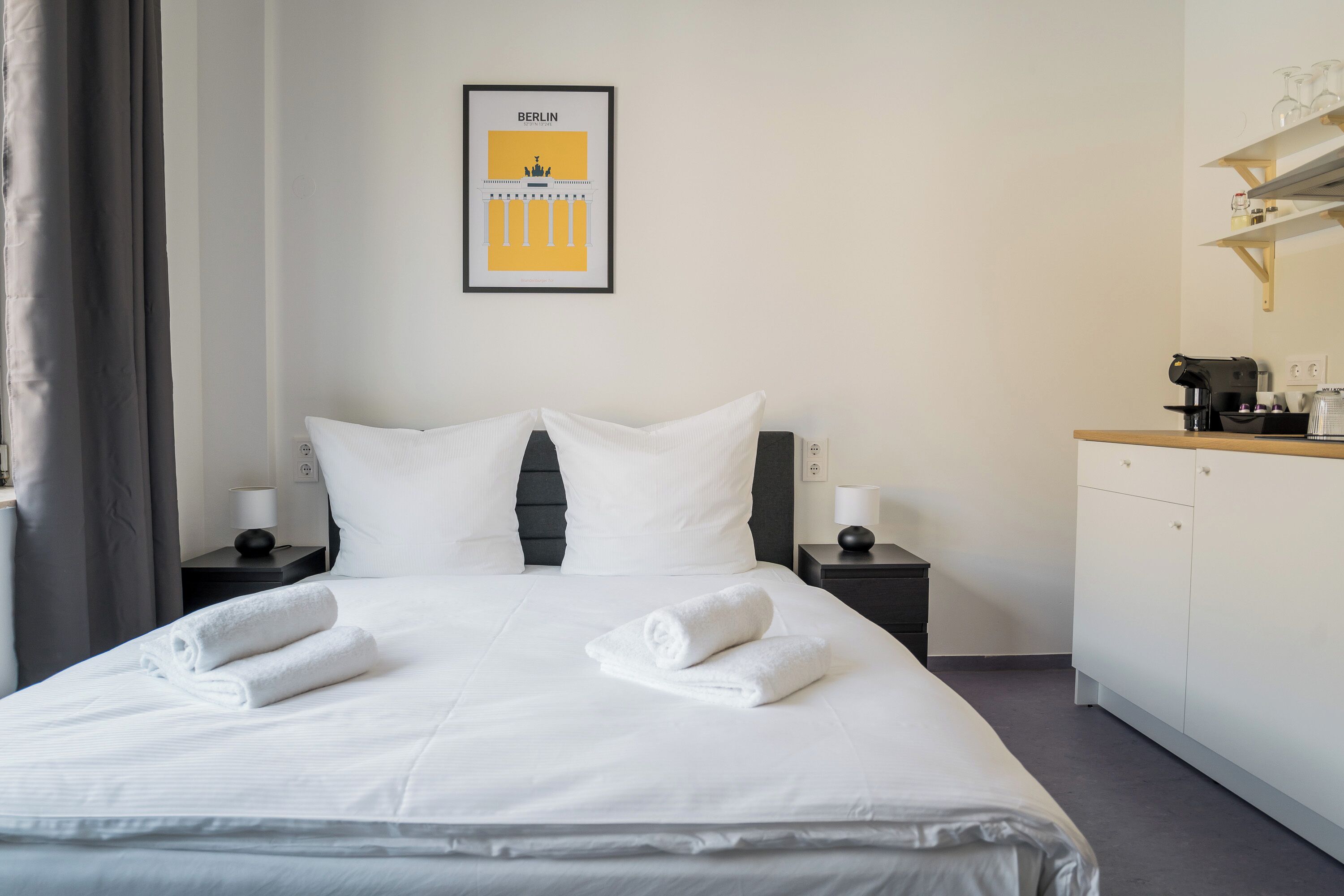 A neatly made bed with white linens and towels, a framed picture of Berlin, a coffee machine, and a small cabinet on a bedside table.