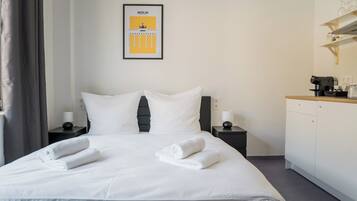A neatly made bed with white linens and towels, a framed picture of Berlin, a coffee machine, and a small cabinet on a bedside table.