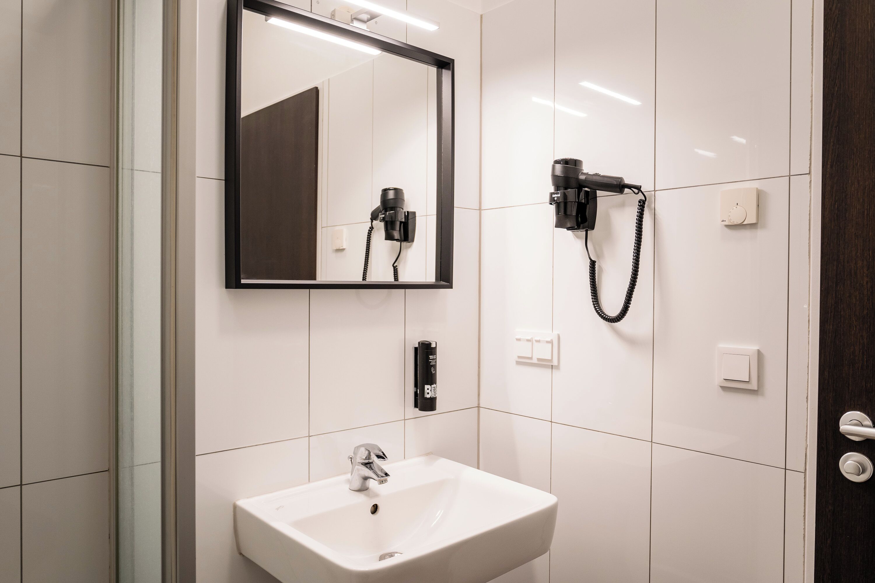 A modern bathroom with a white sink, a large mirror, and a hairdryer mounted on the wall.
