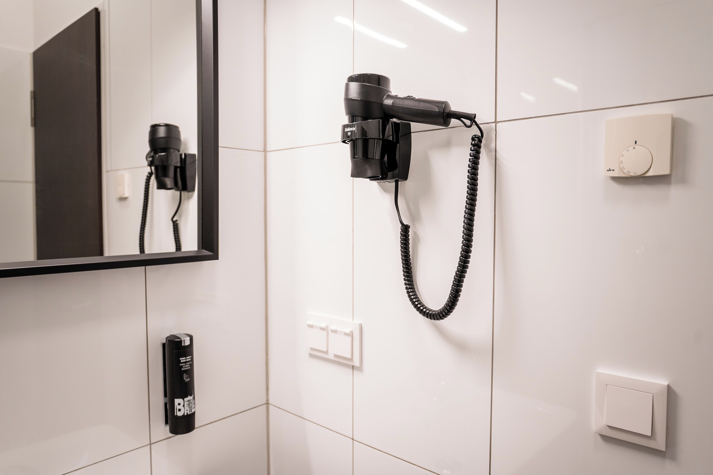 A bathroom with a hairdryer mounted on the wall, a mirror, and a light switch.