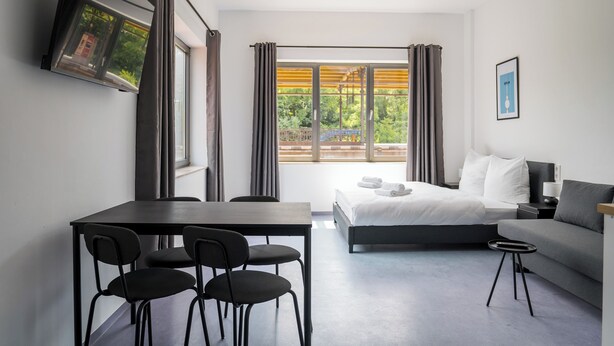 Una moderna camera d'albergo con un letto, una poltrona, un tavolo da pranzo con sedie e un televisore montato a parete.