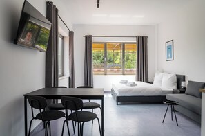 A modern hotel room with a bed, a sofa, a dining table with chairs, and a TV mounted on the wall.