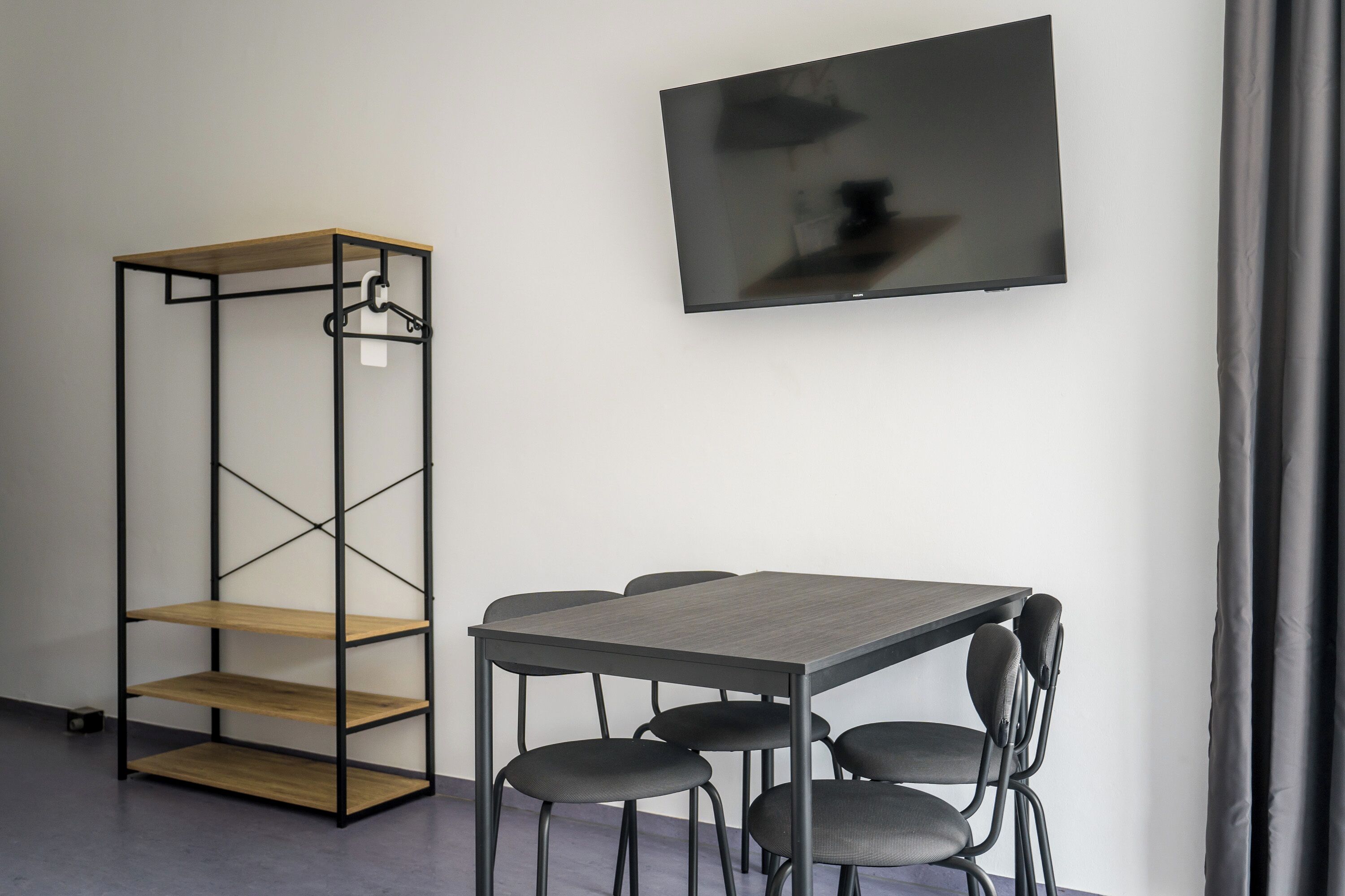 Et moderne værelse med en træreol, et gråt bord med fire stole og et fladskærms-tv ophængt på væggen.
