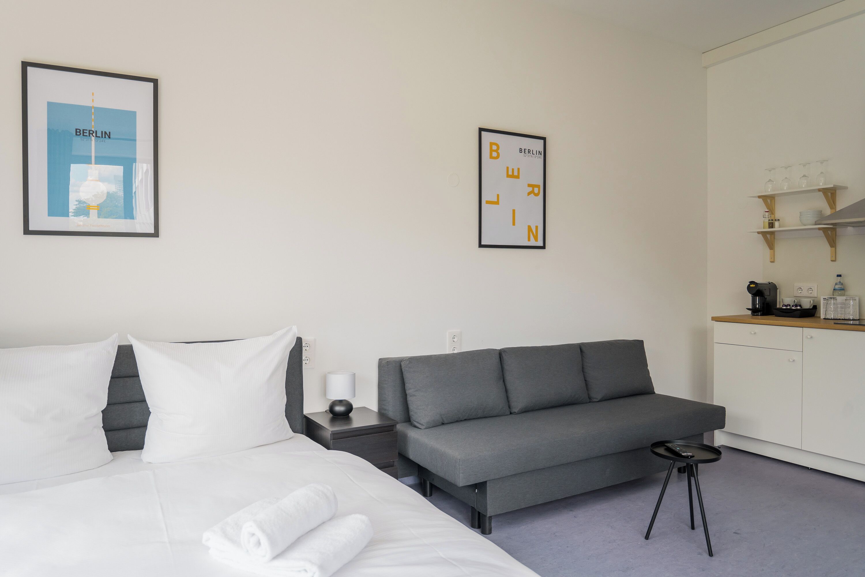 Habitación de hotel con una cama, un sofá gris, una mesita, una mesita de noche, una cafetera y un estante con vasos.