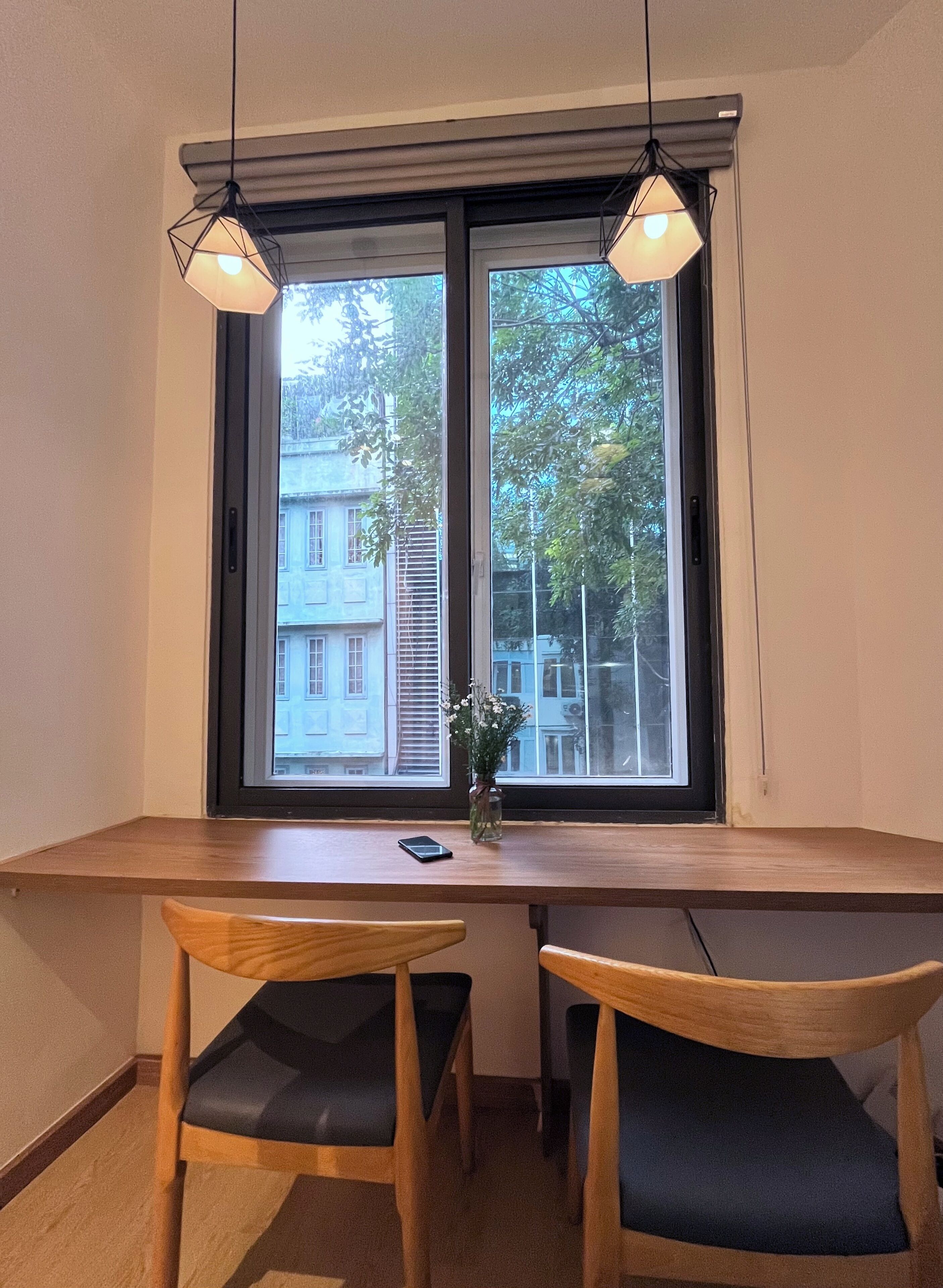 A room with a wooden table and two chairs, a window with blinds, and a vase with flowers on the table.