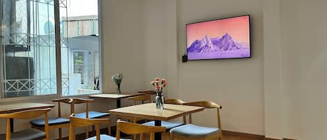 A dining area with wooden tables and chairs, a large window with a view, and a wall-mounted TV displaying a mountain scene.