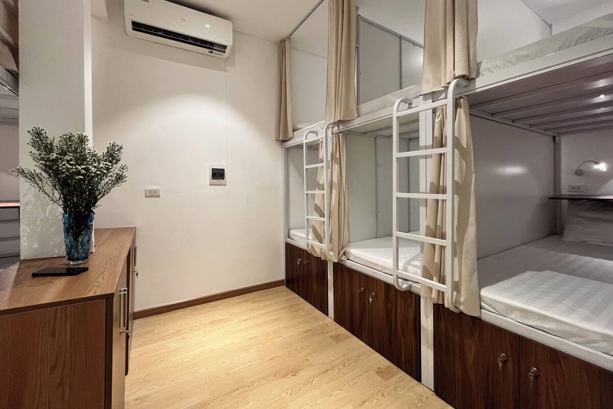 A bunk bed room with a wooden desk, a vase with flowers, an air conditioning unit, and a window with curtains.