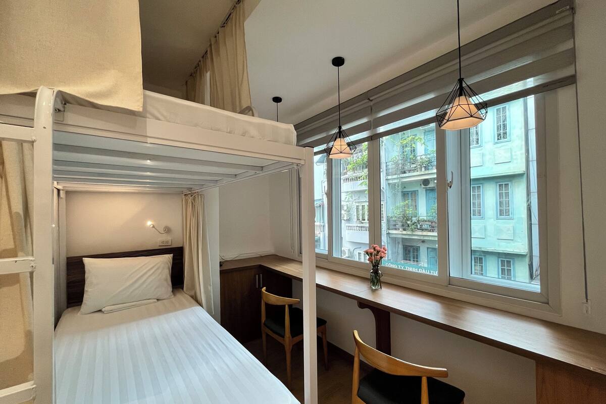 A dorm-style room with bunk beds, a desk, two chairs, a window with a view of buildings, and pendant lights.