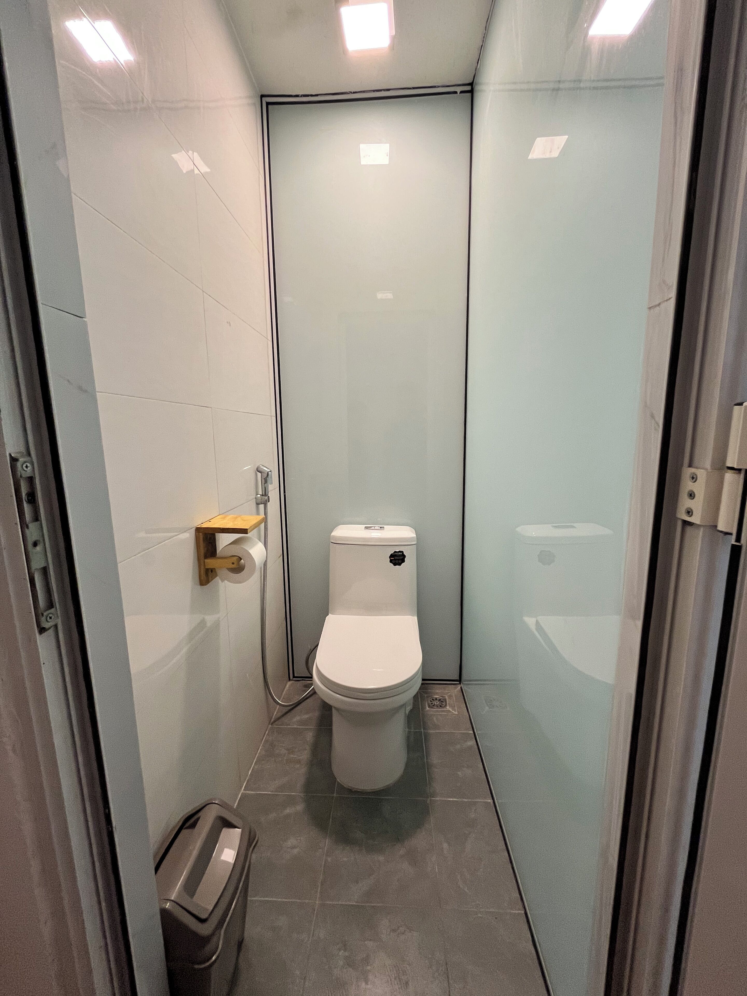 Un baño pequeño y moderno con inodoro, un cesto de basura y mampara de vidrio.