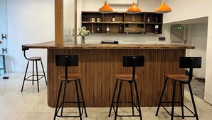 Un área de bar moderna con mostrador de madera, tres taburetes negros y lámparas colgantes naranjas.