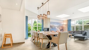 Un salon spacieux avec une grande table à manger, un canapé et un téléviseur. De grandes fenêtres, équipées de rideaux, offrent une vue sur l’extérieur.