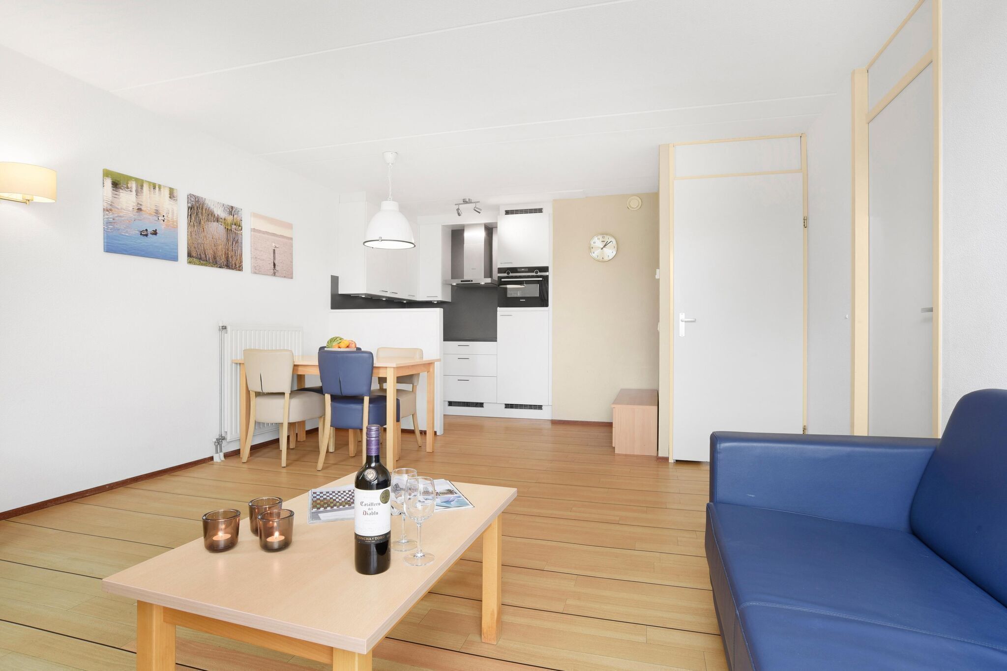 A modern living room with a blue sofa, a wooden coffee table with a bottle of wine and glasses, a dining area with a table and chairs, and a kitchen in the background.