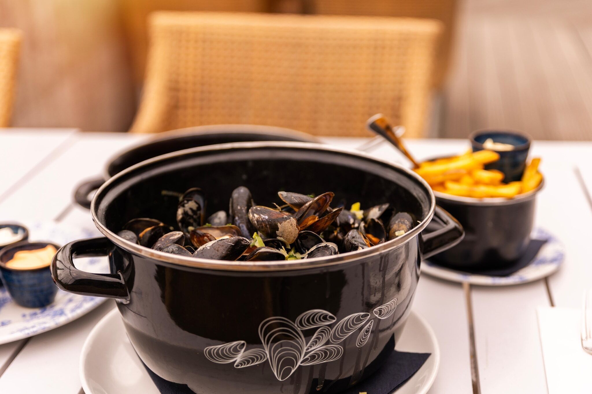 En svart kastrull med musslor står på ett vitt bord, tillsammans med en gaffel och en liten skål med pommes frites.