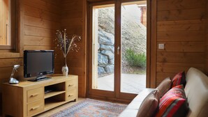 Un interior de madera con un televisor sobre un mueble de madera, un sofá con cojines de colores y vistas a una zona rocosa al exterior a través de un ventanal.