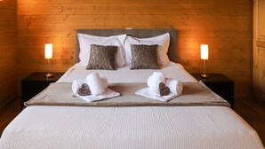 Una cama bien tendida con sábanas blancas y dos toallas con forma de corazón, flanqueada por dos mesitas de noche con lámparas, en una habitación con paredes de madera y techo inclinado.