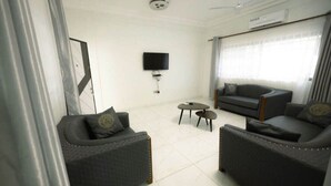 A modern living room with two sofas, a small table, a flat-screen TV mounted on the wall, and a ceiling fan. - DAA DINGBE SUITES (Tamale)