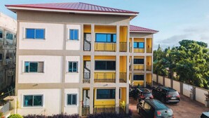 A multi-story building with a red roof, yellow balconies, and a parking area with several cars. - DAA DINGBE SUITES (Tamale)