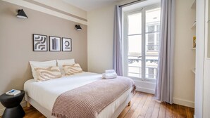 A bedroom with a large bed, a window with curtains, a bedside table, and wall art.