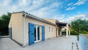 Ein zweistöckiges Haus mit einer gefliesten Terrasse, blauen Fensterläden und einem überdachten Sitzbereich im Freien.