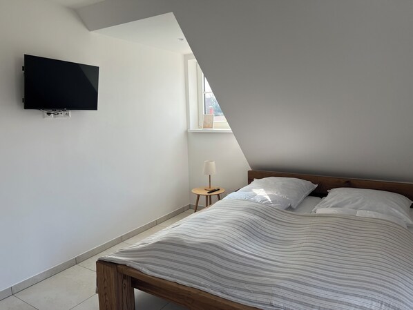A bedroom with a bed, a TV mounted on the wall, a small bedside table with a lamp, and a window with a view of the outside. - Vacation in paradise (Dassow)