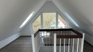 Habitación de desván con techos altos, suelo de madera, una cuna blanca y vistas a árboles y una casa a través de amplios ventanales.